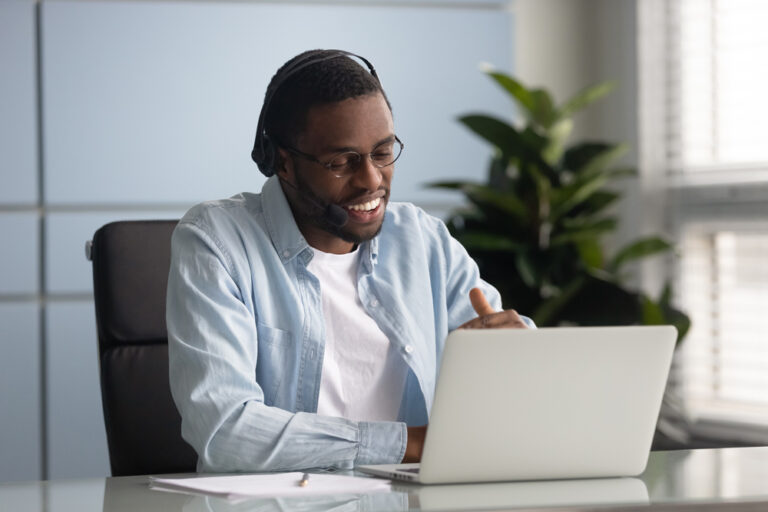 Happy african American male worker wearing headphones speak on video call using laptop, smiling biracial young businessman in earphones have business conference with client at computer online