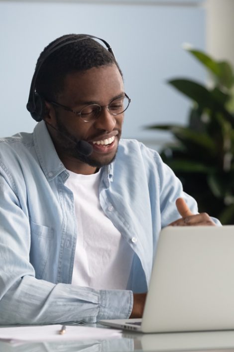 Happy african American male worker wearing headphones speak on video call using laptop, smiling biracial young businessman in earphones have business conference with client at computer online